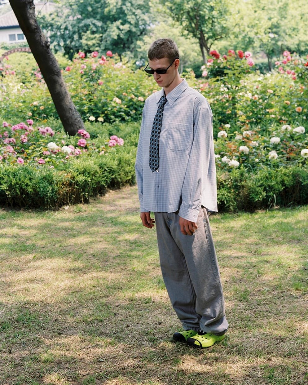 Young man wearing sunglasses, a loose tie, oversized gray pants, and neon green Crocs stands in a rose garden, looking down.