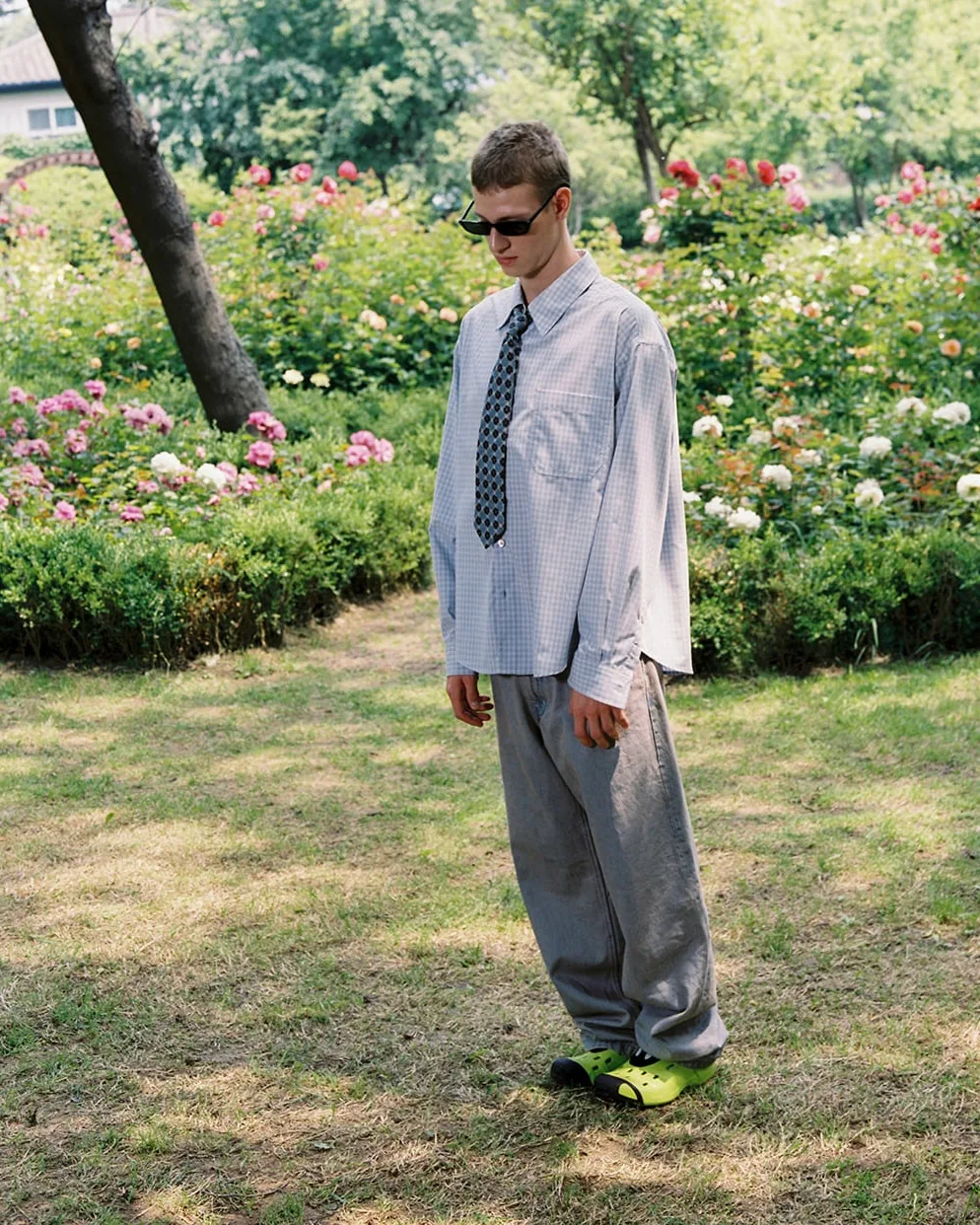 Young man wearing sunglasses, a loose tie, oversized gray pants, and neon green Crocs stands in a rose garden, looking down.