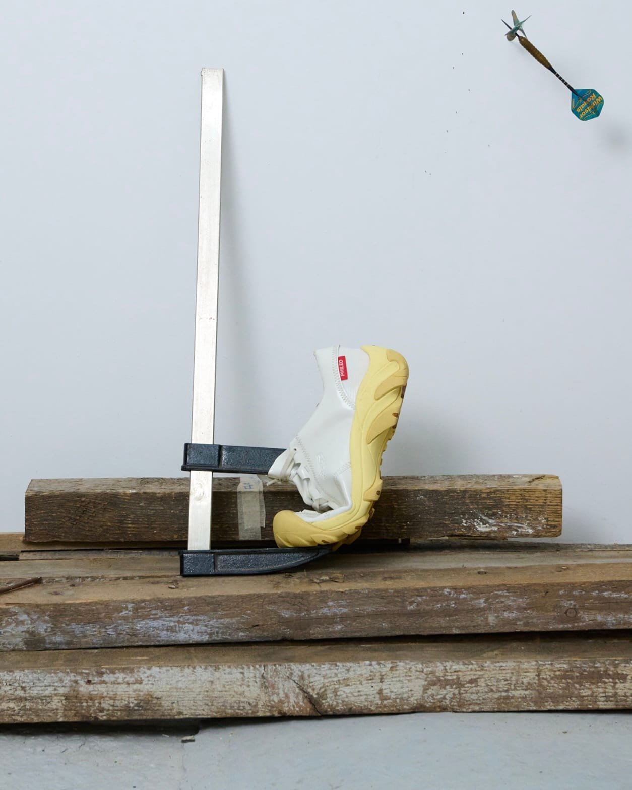 White sneaker with yellow sole clamped to a metal fixture on weathered wooden planks in a workshop.