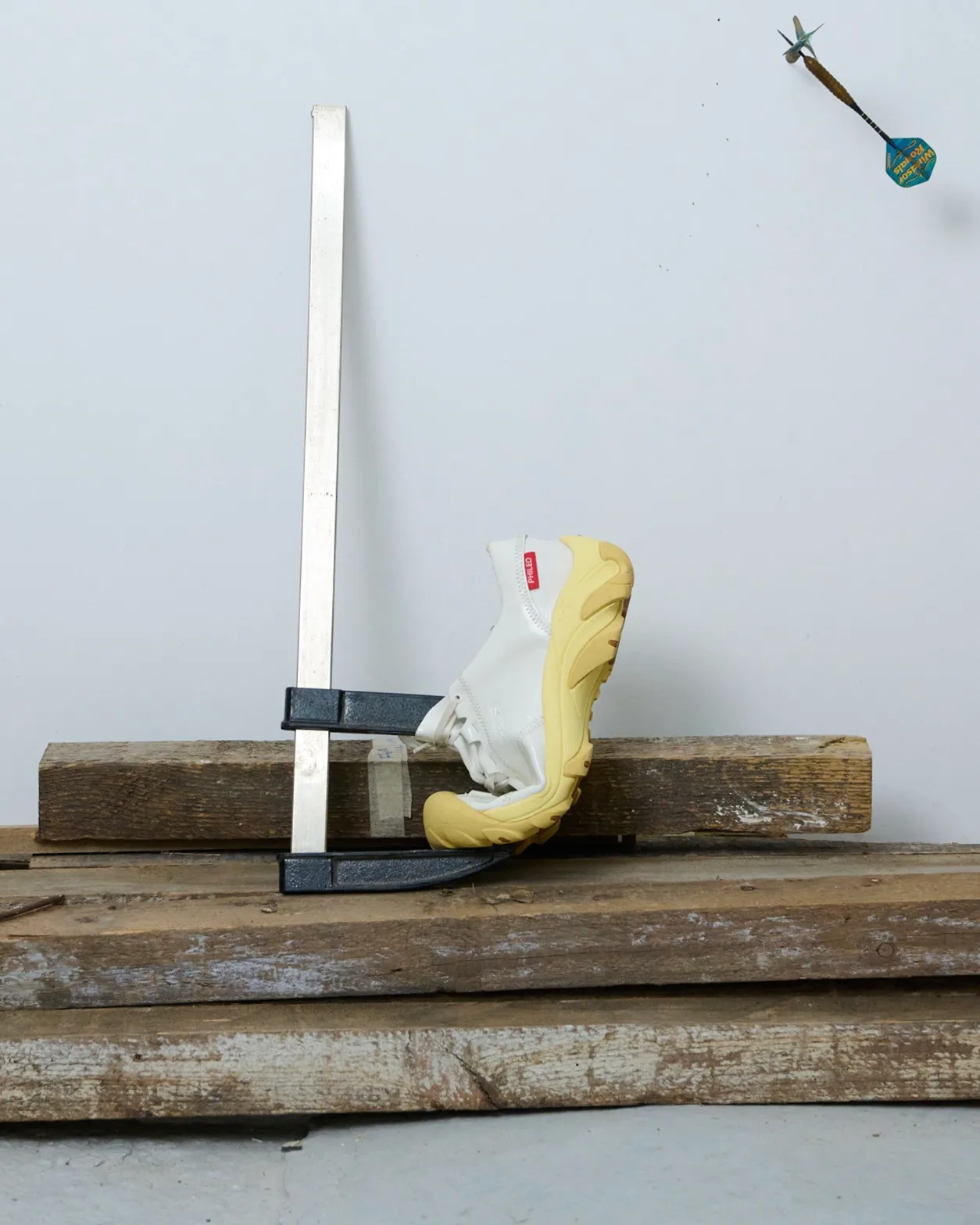 White sneaker with yellow sole clamped to a metal fixture on weathered wooden planks in a workshop.