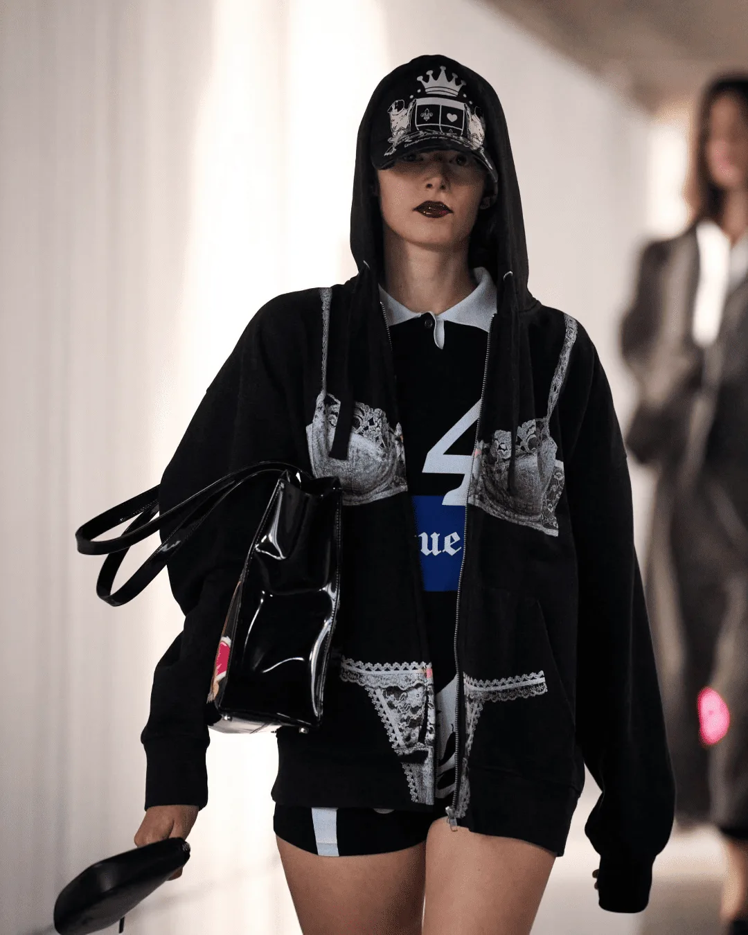Runway model wearing a black hoodie with lace prints and a cap, carrying a glossy black handbag down the catwalk.