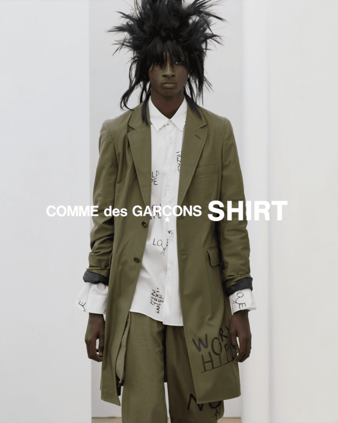 Model wearing an olive-green suit over a white shirt, in a Comme des Garçons SHIRT campaign look with bold, graphic styling and a wild hair silhouette.