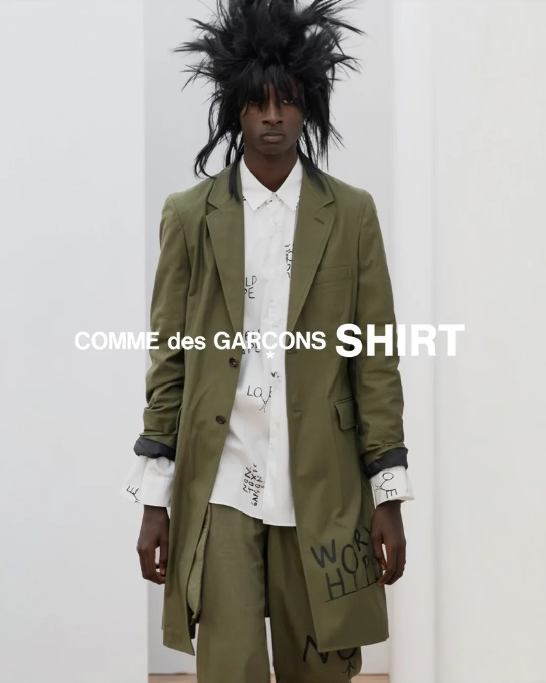 Model wearing an olive-green suit over a white shirt, in a Comme des Garçons SHIRT campaign look with bold, graphic styling and a wild hair silhouette.