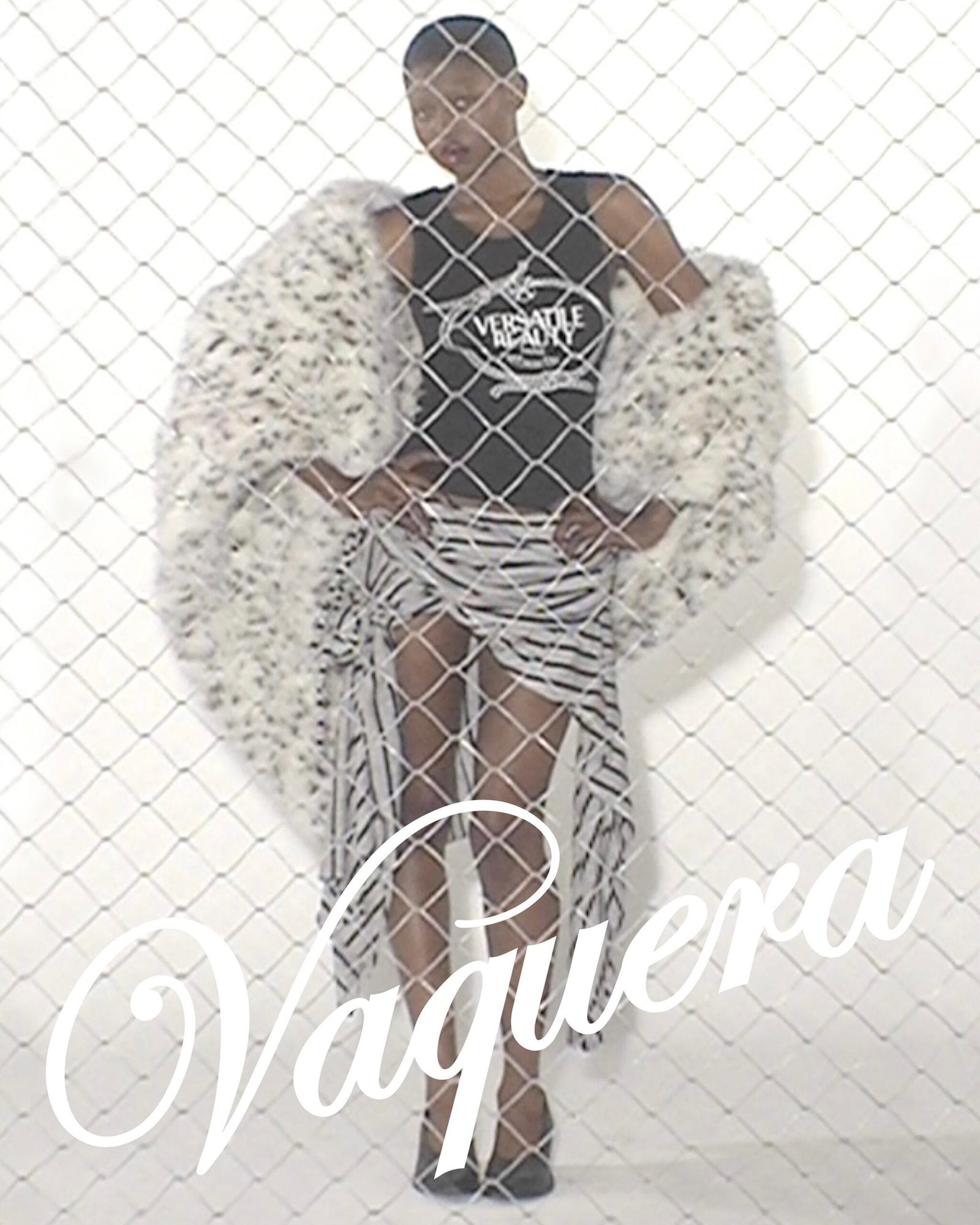 Model behind a chain-link fence wearing a white faux-fur coat, black top, and striped pants.