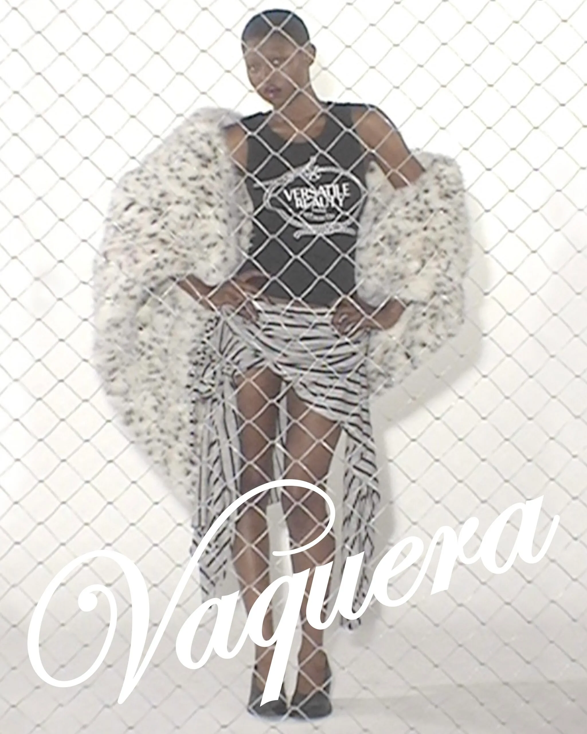 Model behind a chain-link fence wearing a white faux-fur coat, black top, and striped pants.